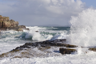 Large white water waves crash through a channel between some rocks, with white foam swirling over the rocks from the largest waves, and sea spray thrown high into the air.