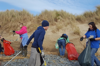 A group of 5 people in brightly coloured clothing, carrying litter pickers and bright red rubbish bags, collecting plastic and other waste from a pebbly area of a beach, in front of some sand dunes.