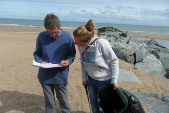 Two women, one with a clipboard and pencil, one with a litter picker and bin bag, both looking at the papers on the clipboard. They are stood on a sandy beach, with some large rocks of a breakwater and the sea behind them.