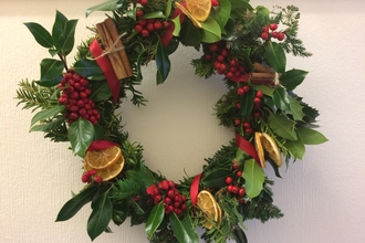 A homemade Christmas wreath with lots of different foliage, leaves, mosses, bright red berries, cinnamon sticks, dried citrus slices, and red ribbon.