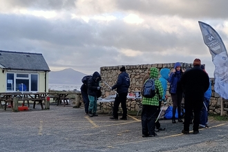 A small group of people stood in a car park talking, with litter picking equipment. Behind them is a large stone wall and fish and chip restaurant. Beyond that the sea is visible in the background, and a small mountain the other side of the water.