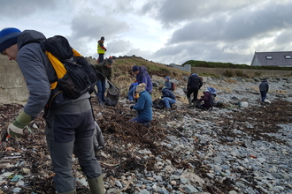 A group of 12 volunteers using litter picking equipment on the strandline at the very top of a pebble beach. Mixed in with the seaweeds some bight colours of plastic rope and other rubbish can be seen, a large white plastic container is lying part way down the beach. The weather is overcast with lots of grey clouds.