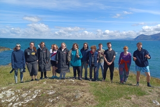 12 young people standing on top of a cliff with the sea behind them all smiling for the camera