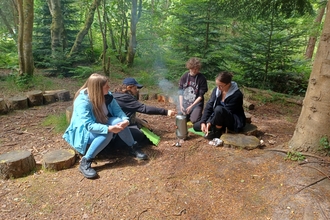 Four trainees lighting and maintaining a kelly kettle whilst sitting on the floor in the woods 