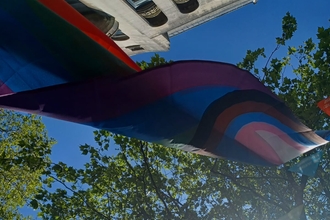 A progress pride flag hangs overhead. above it is a bright blue sky, the tops of city buildings and a large tree with the sunlight cascading through it.