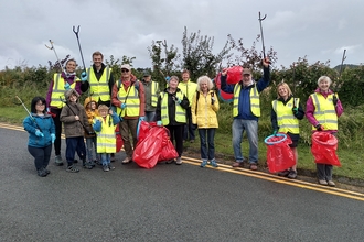  A group of adults and some children, in yellow high visibility vests holding red bin bags and raised up litter pickers, smiling at the camera. They are stood just on the edge of a public road, with a grass verge and hedgerow behind them.