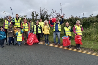  A group of adults and some children, in yellow high visibility vests holding red bin bags and raised up litter pickers, smiling at the camera. They are stood just on the edge of a public road, with a grass verge and hedgerow behind them.