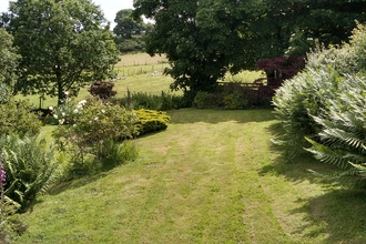 A garden with a grassy lawn between beds of large shrubs and flowering plants. The bottom of the garden is bordered by fencing and trees, with a view through the gaps to fields behind.