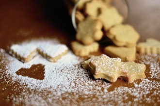 A selection of small shaped biscuits, including hearts, stars and flowers are spilled out of a container onto a table. Some biscuits are dusted with icing sugar, and a heart shape gap in the icing sugar is left where one has been moved afterwards.