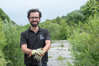 A white man with short dark hair and beard. He is wearing a black polo shirt with the NWWT logo, glasses and gardening gloves, stood smiling at the camera while leaning on a gardening implement. Behind him there is a gravely clearing surrounded by large bushes and trees.