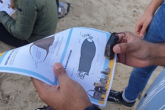 A man holds a shark eggcase, and an ID guide, matching the shape and appearance to the picture. On the beach behind another person sits in the sand doing the same thing to identify a different eggcase.