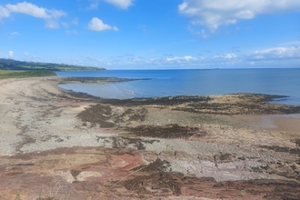 A rocky coastline, with pebble and sand beach. The sea is like glass, calm, flat and mid blue. The sky is bright with rows of fluffy white clouds 