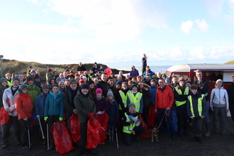 A large crowd is gathered in a car park with a beach and the sea behind them. Some are wearing high vis vests, most are holding litter pickers and red or blue bin bags.