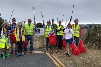 A small group of adults and one child, dressed in high vis and holding red bin bags and litterpickers. They are stood at the corner of a road with a field behind them, and a sign on the boundary gate with the North Wales Wildlife Trust logo. Text reads ' Caring for the natural environment of North Wales. Protecting wildlife for the future and for all to enjoy', in Welsh and English.