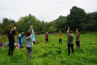 A group of people stood in a circle in a field. They have their arms raised as they move through yoga poses.