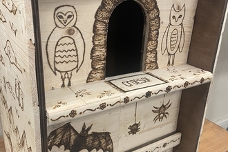 A hand-crafted wooden owl box intricately decorated with pyrography art. The designs include owls, bats, snails, spiders, and floral patterns, all burned into the wood. The box features a central opening for nesting and has detailed artwork inspired by children's drawings.