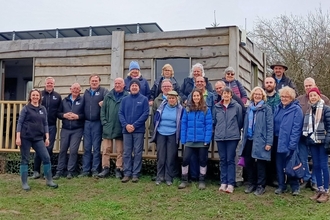 A group of 22 volunteers and staff, wearing mainly blue fleeces or waterproofs, stood on the grass outside a wooden single level building. 