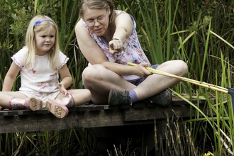 A young girl and a woman sat cross-legged on a wooden platform, the woman is holding fishing nets and pointing outwards towards the camera. They are surrounded by reeds and other vegetation that grows around water.