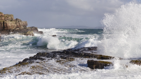 Large white water waves crash through a channel between some rocks, with white foam swirling over the rocks from the largest waves, and sea spray thrown high into the air.