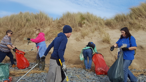 A group of 5 people in brightly coloured clothing, carrying litter pickers and bright red rubbish bags, collecting plastic and other waste from a pebbly area of a beach, in front of some sand dunes.