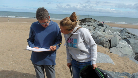 Two women, one with a clipboard and pencil, one with a litter picker and bin bag, both looking at the papers on the clipboard. They are stood on a sandy beach, with some large rocks of a breakwater and the sea behind them.