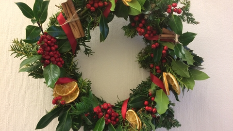 A homemade Christmas wreath with lots of different foliage, leaves, mosses, bright red berries, cinnamon sticks, dried citrus slices, and red ribbon.