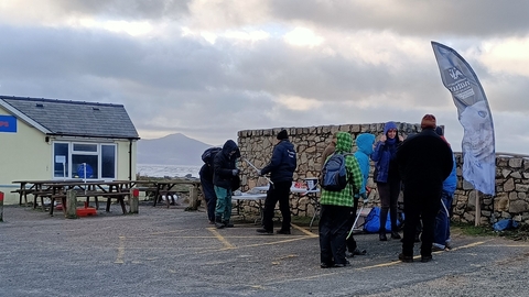 A small group of people stood in a car park talking, with litter picking equipment. Behind them is a large stone wall and fish and chip restaurant. Beyond that the sea is visible in the background, and a small mountain the other side of the water.