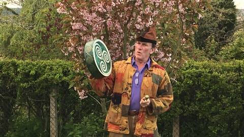 Andy Harrop Smith, Storyteller. Andy, wearing a patchwork coat in autumn colours, holding a hand drum with Celtic triskelion pattern. He is stood in front of a hedge and small tree with lots of pale pink flowers.