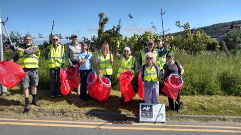 A group of 10 people smiling at the camera on a road verge. They are wearing high vis, holding up litter pickers and red bin bags. On has a sign with the North Wales Wildlife Trust logo on it.