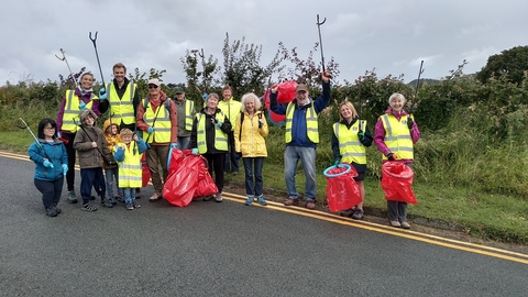  A group of adults and some children, in yellow high visibility vests holding red bin bags and raised up litter pickers, smiling at the camera. They are stood just on the edge of a public road, with a grass verge and hedgerow behind them.