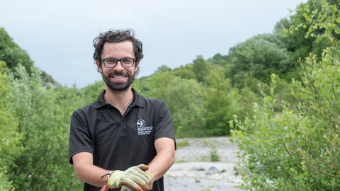 A white man with short dark hair and beard. He is wearing a black polo shirt with the NWWT logo, glasses and gardening gloves, stood smiling at the camera while leaning on a gardening implement. Behind him there is a gravely clearing surrounded by large bushes and trees.