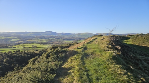 Looking out from a grass ridge across a small valley, farm fields and scrub land nearby with hills in the distance meeting a pale blue sky.