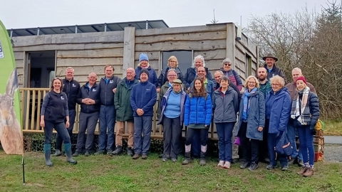 A group of 22 volunteers and staff, wearing mainly blue fleeces or waterproofs, stood on the grass outside a wooden single level building. 