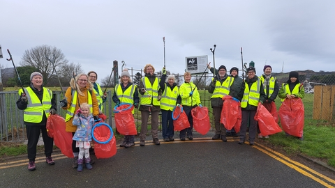 A dozen adults in high vis vests, and a young child. All are holding up litter pickers and red bin bags, while smiling. They are at the corner of a road and a park, one person holds a sign with the NWWT logo.