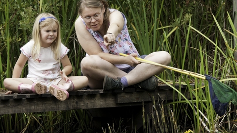 A young girl and a woman sat cross-legged on a wooden platform, the woman is holding fishing nets and pointing outwards towards the camera. They are surrounded by reeds and other vegetation that grows around water.