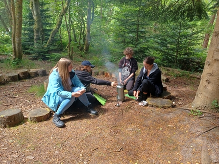 Four trainees lighting and maintaining a kelly kettle whilst sitting on the floor in the woods 