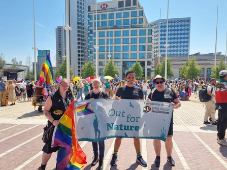 4 people with rainbow flags and face paint stand in a crowded city square. They are holding up a banner that reads: out for nature.