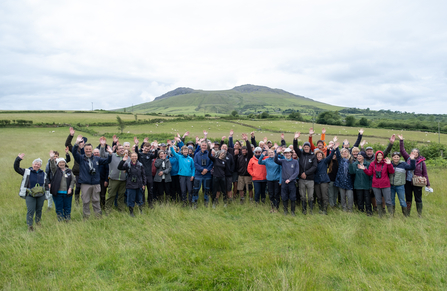 A crowd of people smile and wave at the camera in front of farmland