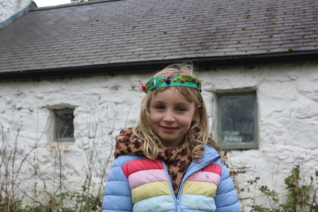 A young girl with blonde hair smiles while wearing a handmade nature crown adorned with flowers, leaves, and seeds. She is dressed warmly in a multicolored striped puffer jacket and a leopard-print scarf. Behind her is a rustic white stone building with a slate roof and small, old windows, surrounded by bare branches and greenery, suggesting an autumn or early winter setting.