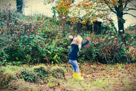 A blurred image of a child mid-jump, dressed in a navy sweater, colorful leggings, and bright yellow rain boots, playing in an outdoor setting. The background features dense greenery with wild plants, shrubs, and a tree with autumn-colored leaves. The scene captures the energy and joy of outdoor play in a natural, untamed environment.