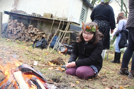 A young girl wearing glasses and a nature-inspired crown made of leaves and feathers sits by a small campfire outdoors. She is dressed warmly in a black coat and patterned leggings, with a calm expression. In the background, there are stacked firewood, backpacks, and a few other people engaged in activities, some also wearing nature crowns. The setting appears to be a rustic, outdoor environment with scattered autumn leaves and natural elements.