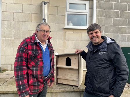 Neil Dunsire and Robin Lomas standing outside in front of a light-colored brick wall, smiling at the camera. They are showcasing a plain wooden owl box, designed for conservation purposes, which Robin’s company, The Owl Box, created. Robin is wearing a red and black plaid jacket, while Neil is wearing a North Wales Wildlife Trust jacket. The owl box sits on a small ledge beside them.