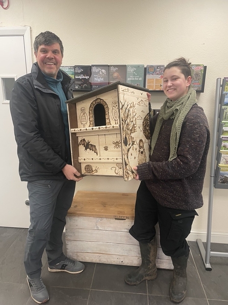 Two individuals, Neil Dunsire and Ginny West, standing indoors and holding a beautifully pyrography-decorated owl box. Both are smiling, showcasing the completed project. The background features posters and informational displays related to nature and wildlife conservation.