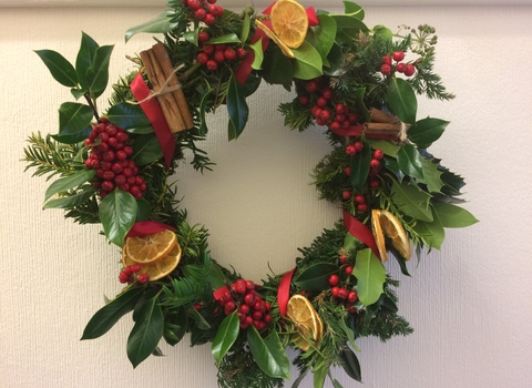 A homemade Christmas wreath with lots of different foliage, leaves, mosses, bright red berries, cinnamon sticks, dried citrus slices, and red ribbon.