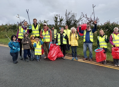  A group of adults and some children, in yellow high visibility vests holding red bin bags and raised up litter pickers, smiling at the camera. They are stood just on the edge of a public road, with a grass verge and hedgerow behind them.